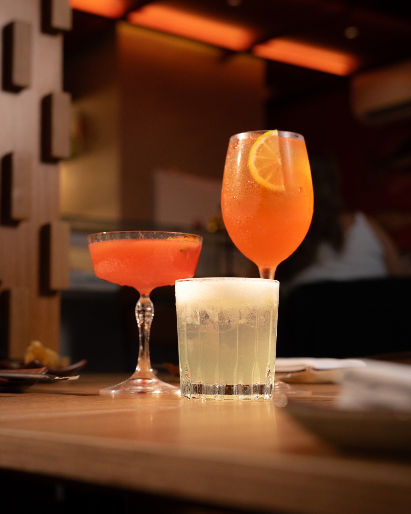 Trio de drinks autorais em taças diferentes sobre balcão de madeira com iluminação âmbar ao fundo