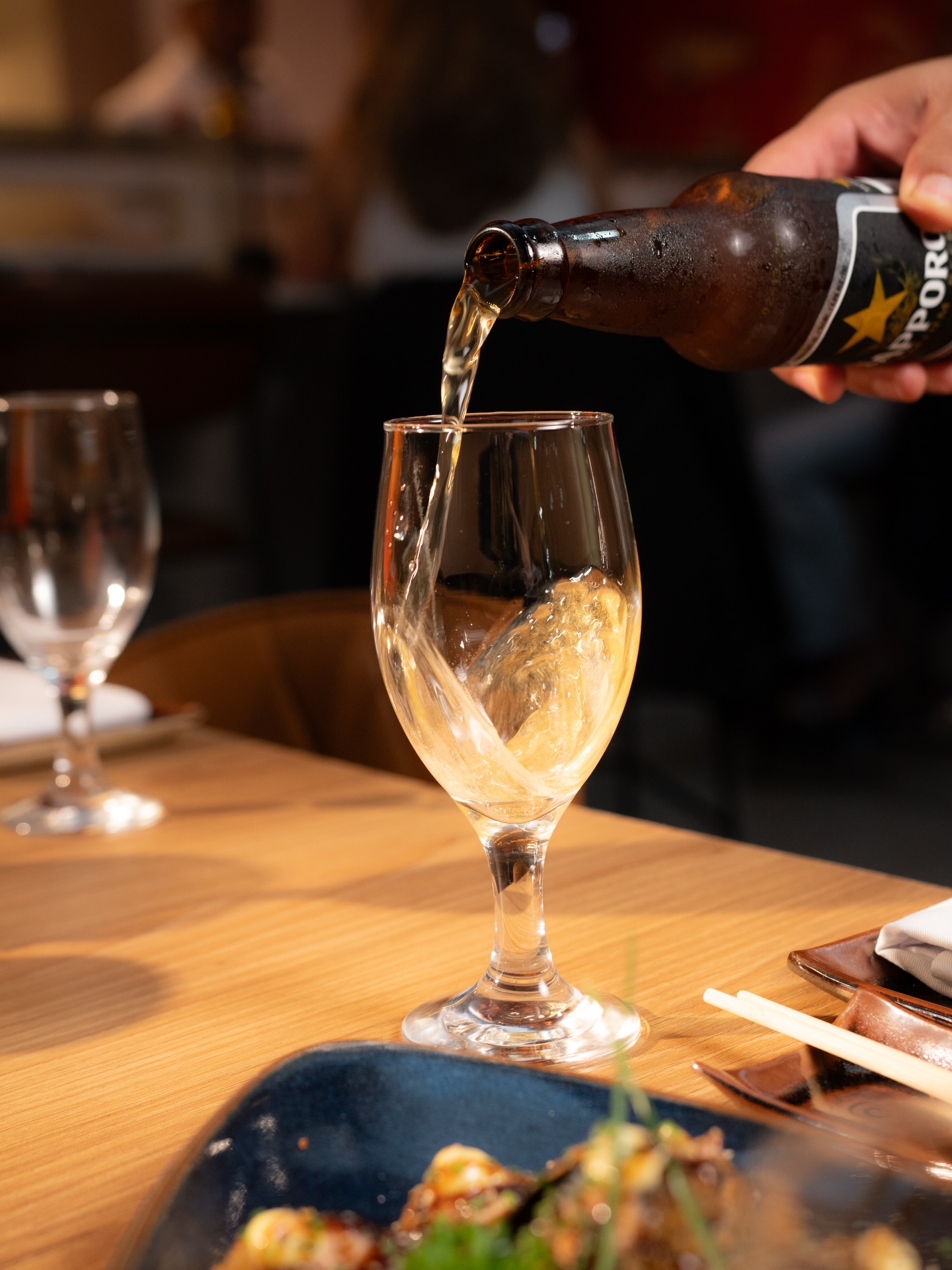 Cerveja Sapporo sendo servida em taça sobre mesa de madeira com prato em segundo plano