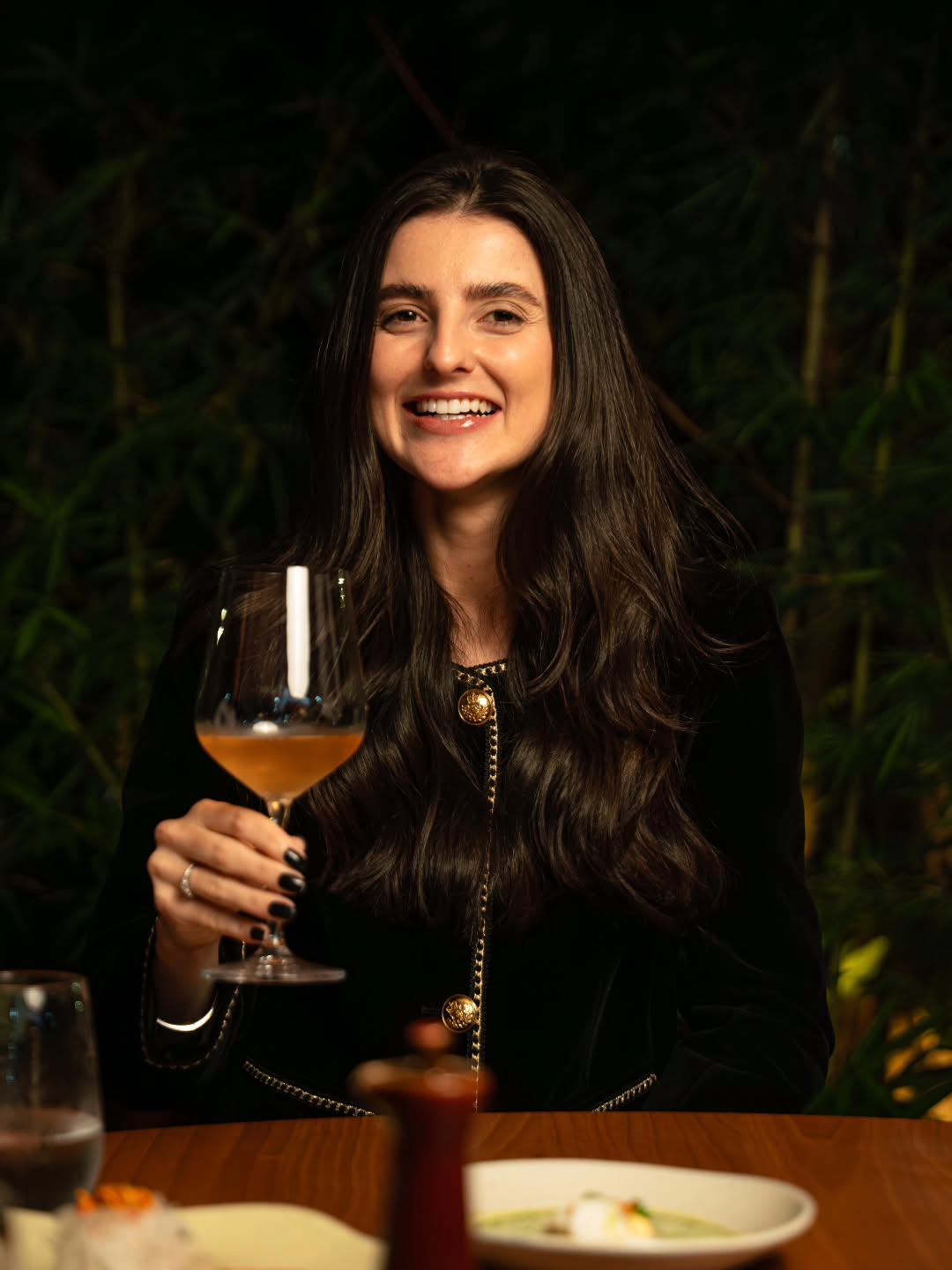 Convidada sorrindo com taça de vinho branco no salão intimista do Shakkai, com bambus ao fundo