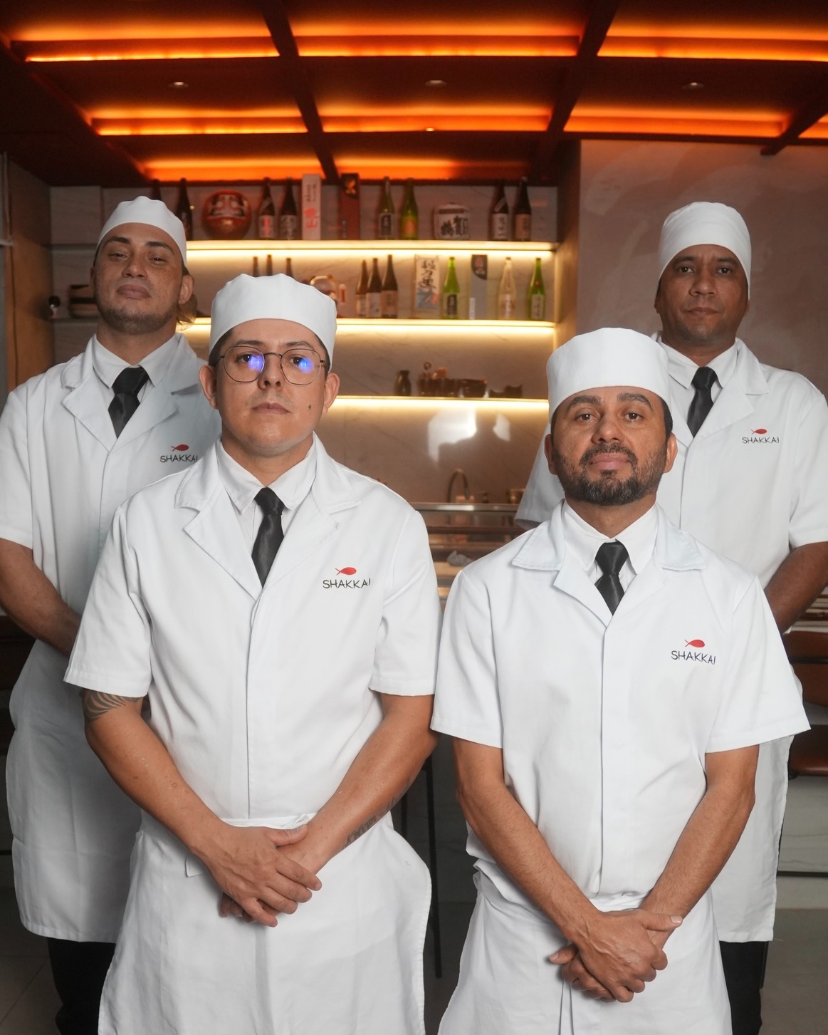 Quatro sushimen uniformizados posicionados diante do balcão de sakes iluminado no salão do Shakkai