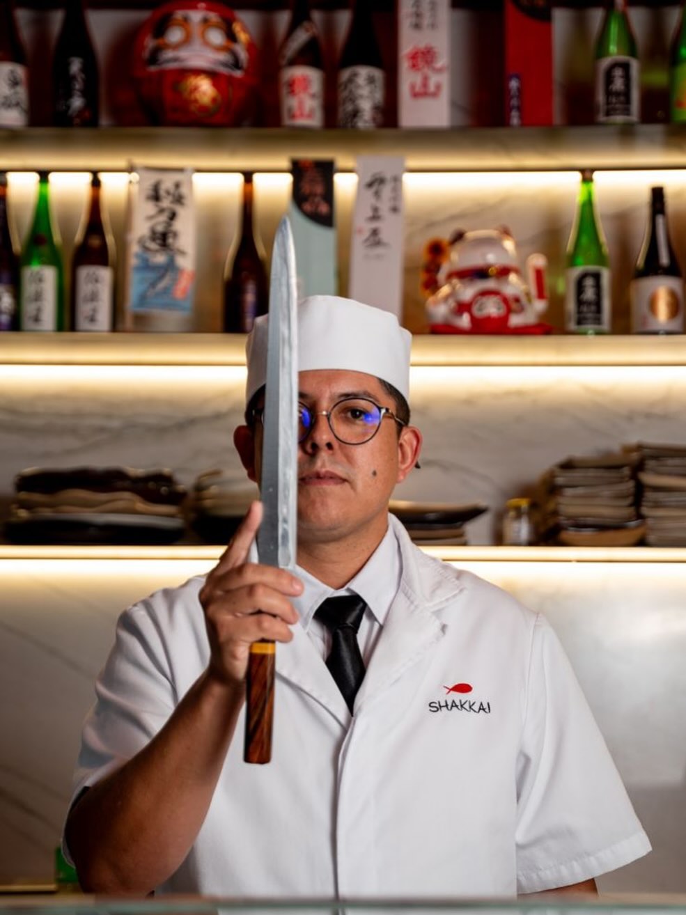 Chef Shakkai em uniforme branco segurando faca japonesa diante do balcão de sakes iluminado