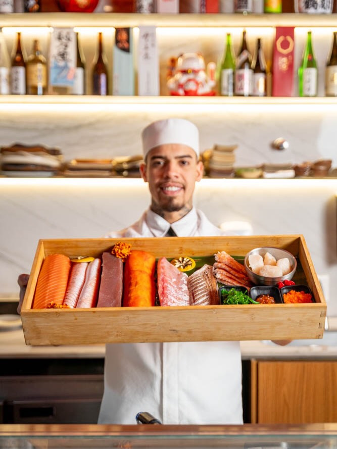 Sushiman uniformizado sorrindo enquanto apresenta bandeja de madeira com cortes de salmão, atum, camarão, vieira e ovas diante do balcão de sakes