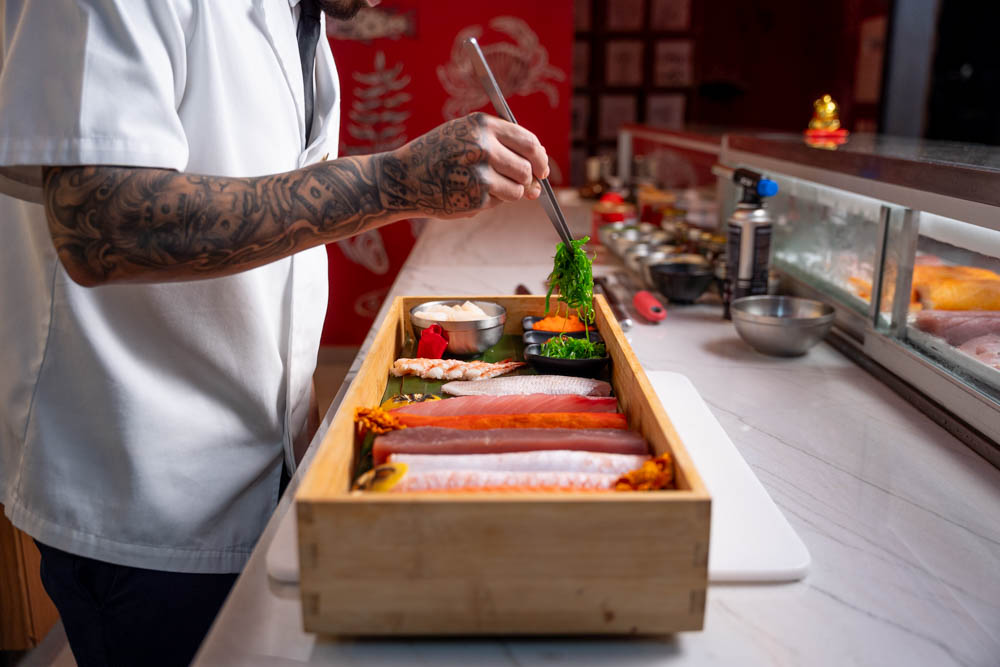 Chef tatuado montando bandeja de peixes no balcão com parede vermelha ilustrada ao fundo