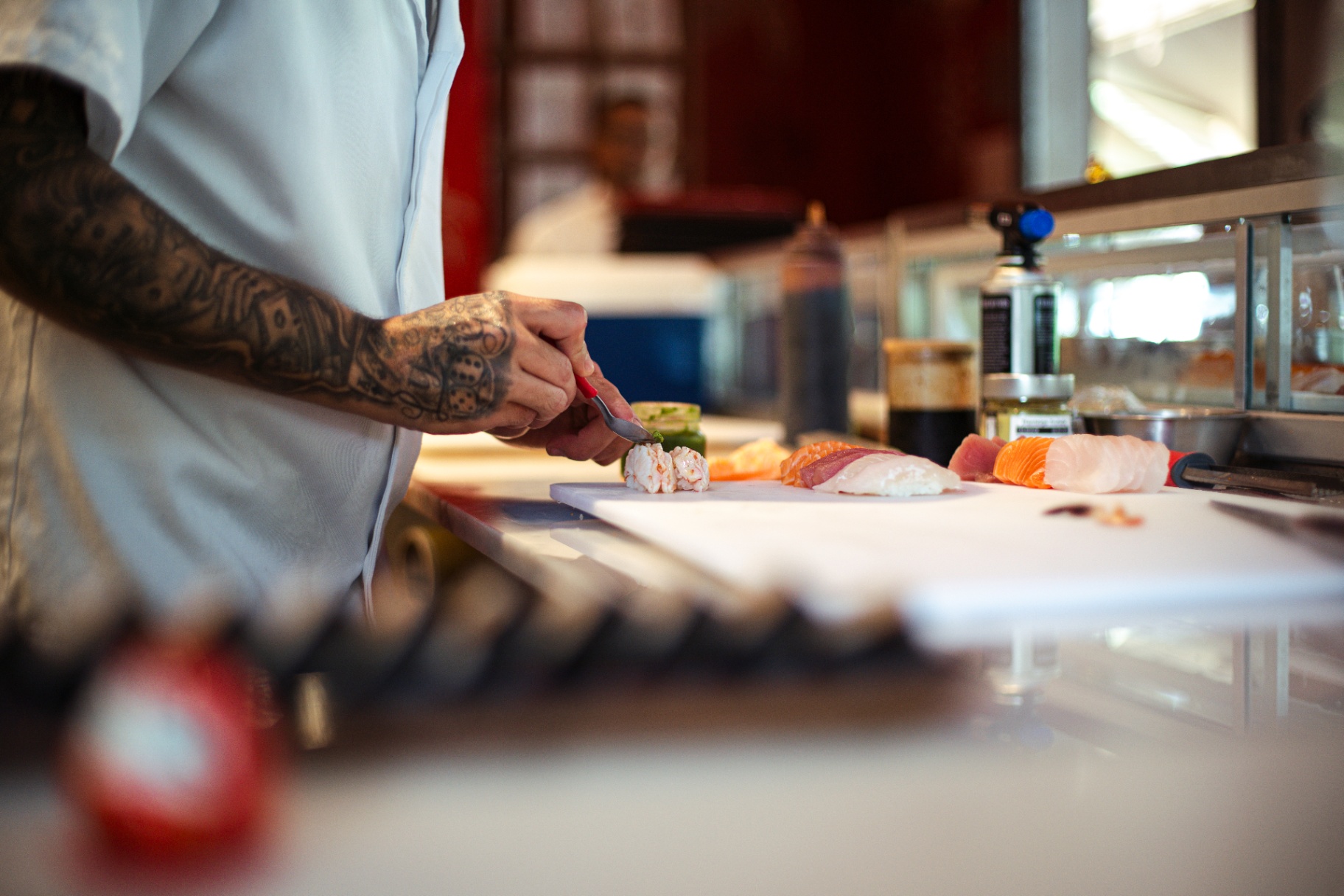Chef de braço tatuado finalizando nigiri no balcão com mise en place de salmão e atum em primeiro plano desfocado