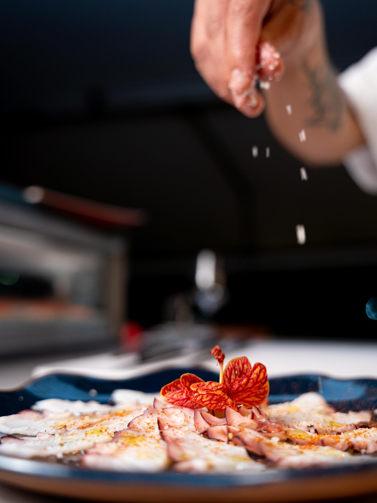 Mão do chef finalizando com flor de sal um carpaccio de polvo em prato azul com flor comestível vermelha