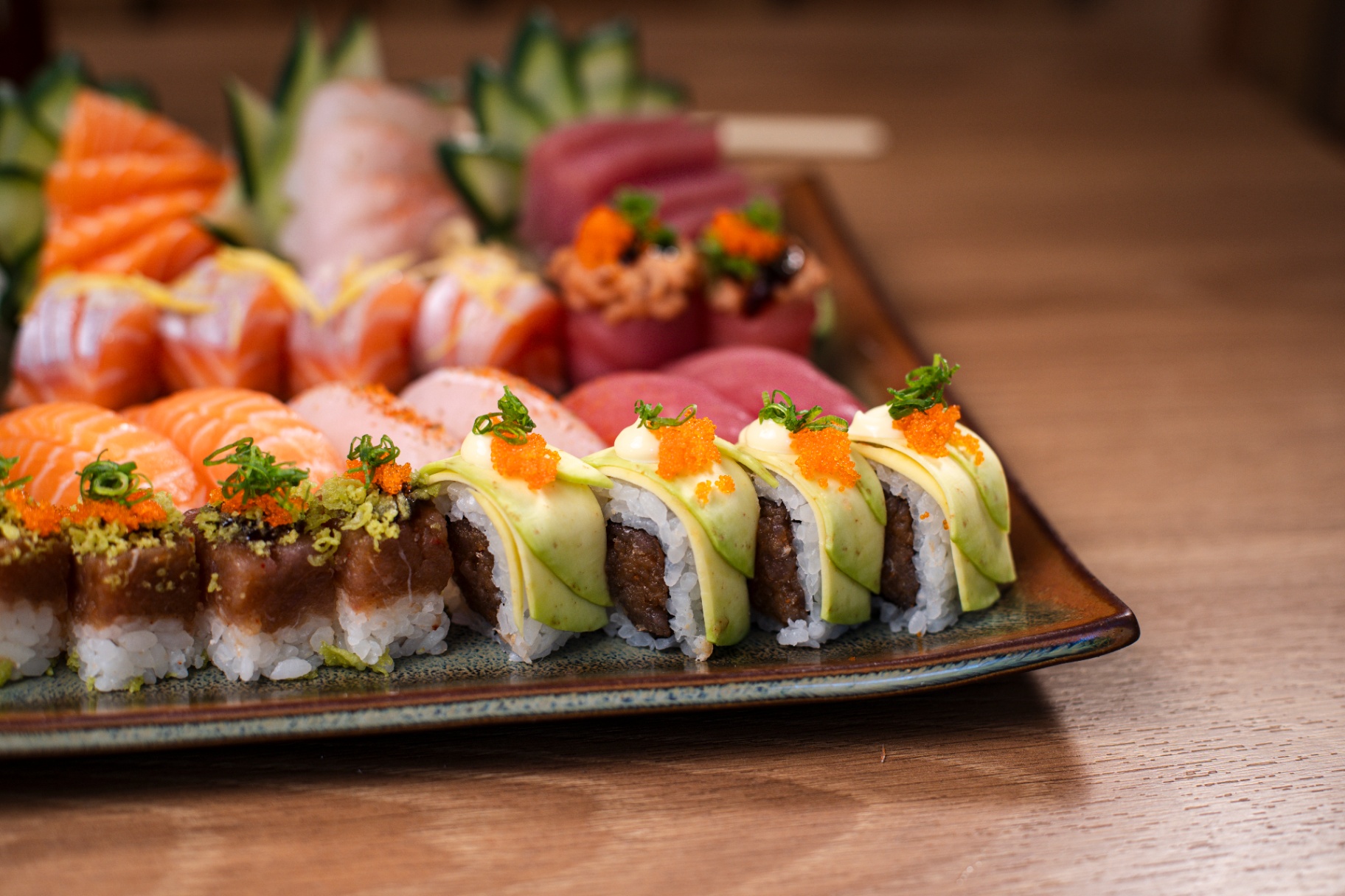 Bandeja horizontal com sashimi de salmão e atum ao fundo e hot roll com avocado e ovas em primeiro plano